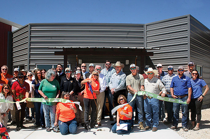 A group of people are lined up facing the camera and have just apparently cut through a large ribbon held in front of them with the new, gray metal Kerr County Animal Control facility in the background.